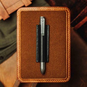 May include: A black leather pen holder with a silver pen, presented on a brown rectangular case with a stitched border. The pen features a silver clip with a small design. The case has a textured, felt-like surface.