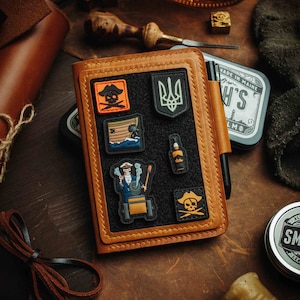 May include: A tan leather notebook with a black interior and various pirate-themed patches. A pen is held in a leather loop. Other leather items and tools surround the notebook, suggesting a craft or workshop setting.