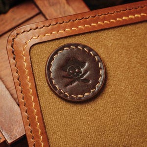 May include: Close-up of a brown leather wallet with a stitched border and a circular emblem. The emblem features a skull and crossbones wearing a pirate hat. The wallet has a tan interior.