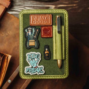 Puede incluir: Un portalápices de cuero verde con un bolígrafo y varios parches. Los parches incluyen las palabras "PIRATE GOODS" y "EDC FREAK". Otros parches presentan un esqueleto, una botella y un logotipo. El bolígrafo es negro y dorado.