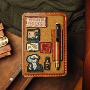 Op de afbeelding: Een bruin leren notitieboek met een penhouder en verschillende patches. Patches bevatten de tekst "PIRATE GOODS", een schip, een kaart, een schedel en een fles. De pen is bruin met een zwarte clip en een schedelontwerp.