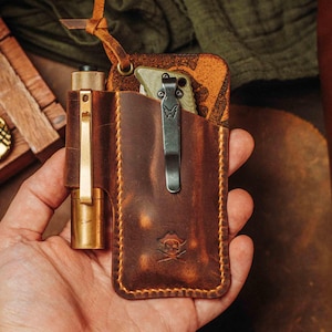 May include: A brown leather pocket organizer with a brass flashlight and a folding knife. The organizer has a stitched edge and a skull and crossbones emblem. A leather strap is attached to the top.