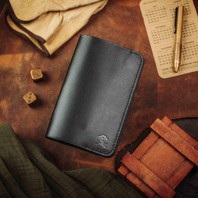 May include: A black leather wallet with a skull and crossbones logo. The wallet is closed and lying on a brown leather surface.