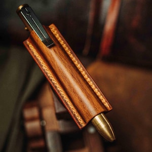 Puede incluir: Funda de cuero marrón con costuras detalladas, que contiene un bolígrafo de latón con un clip plateado. El bolígrafo es parcialmente visible, lo que se suma al diseño. La funda es de color marrón intenso, y las costuras son de un tono más claro.