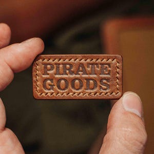 May include: A rectangular, brown leather tag with the words "PIRATE GOODS" embossed in the center. The tag has a stitched border and is held between two fingers. The background is blurred, suggesting a close-up shot.