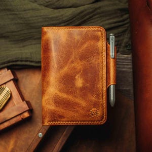 Può includere: Un taccuino in pelle marrone con un portapenne. Il taccuino ha una texture invecchiata e un bordo cucito. Una penna di metallo è nel supporto. Un logo con teschio e ossa incrociate è nell'angolo in basso a destra.