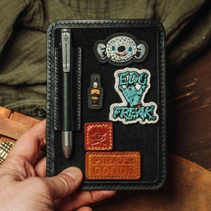 Puede incluir: Organizador EDC de cuero negro con un bolígrafo, parches y un borde cosido. Los parches incluyen un personaje de dibujos animados, una botella y las palabras "EDC FREAK". El parche inferior dice "PIRATE GOODS".