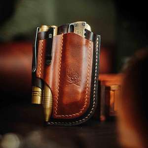 May include: A brown leather pocket organizer featuring a stamped skull and crossbones emblem. The organizer holds a brass pen with a skull clip and a brass folding tool with the letters Br visible on the handle. The leather has dark edges.