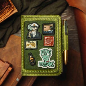 May include: Green leather notebook with a pen holder and various patches. Patches feature a skull in a captain's hat, a ship, a map, a skull and crossbones, a bottle, and the words "EDC FREAK".