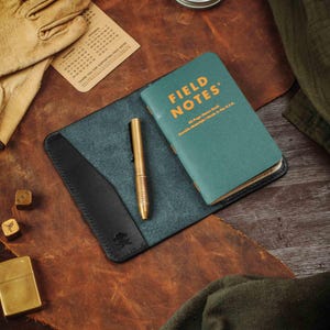 May include: A black leather notebook cover with a blue interior and a pen. The cover has a pocket for a small notebook. The notebook is a Field Notes brand notebook with the text "FIELD NOTES" in orange on a green cover. The text "48-Page Memo Book" and "Pocket Notebook / Made in the U.S.A." is printed in white on the cover.