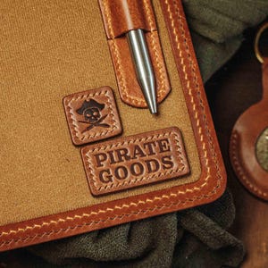 May include: Close-up of a brown leather notebook with a pen holder and a silver pen. The notebook has a stitched border and two leather patches with a skull and crossbones and the words "PIRATE GOODS".