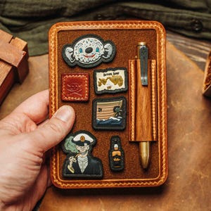 Puede incluir: Un cuaderno de cuero marrón con un bolígrafo de madera y varios parches. Los parches incluyen una calavera, un mapa y un personaje de dibujos animados. El bolígrafo es de madera y metal. El cuaderno lo sostiene una persona.