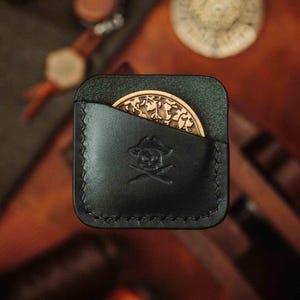 K&ouml;nnte beinhalten: Eine dunkelgr&uuml;ne Lederm&uuml;nzb&ouml;rse mit Totenkopf- und Knochenkreuz-Design. Die B&ouml;rse hat eine Klappe, die sich &ouml;ffnet und eine Tasche f&uuml;r eine M&uuml;nze enth&uuml;llt.