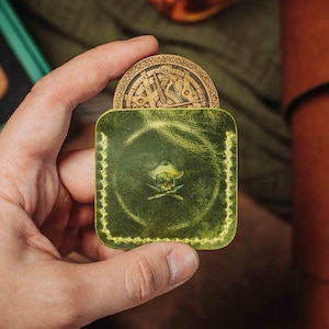 Könnte beinhalten: Ein handgefertigtes, grünes Lederportemonnaie mit Piratenmotiv, das eine goldfarbene Münze enthält. Das Portemonnaie hat ein Totenkopf-Emblem und wird in einer Hand gehalten.