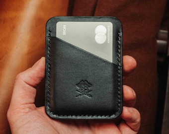May include: A black leather card holder with a stitched edge and a skull and crossbones logo. The holder has a pocket for a credit card, which is visible in the image. The card is a silver Mastercard with the word "IRON" printed on it.