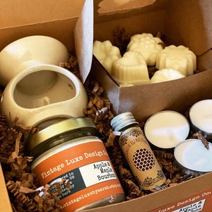 May include: A gift box filled with a white ceramic wax warmer, a jar of apple and maple bourbon scented wax melts, a small bottle of essential oil, and three white tea lights. The box is lined with brown shredded paper.