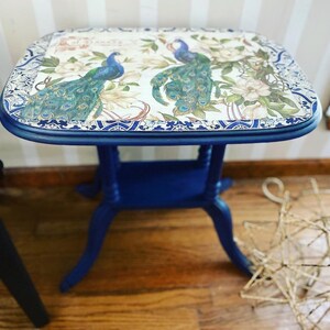 Pode incluir: Uma mesa auxiliar azul com um tampo branco com um design de pav&atilde;o. A mesa tem uma borda decorativa em torno do topo e tr&ecirc;s pernas.