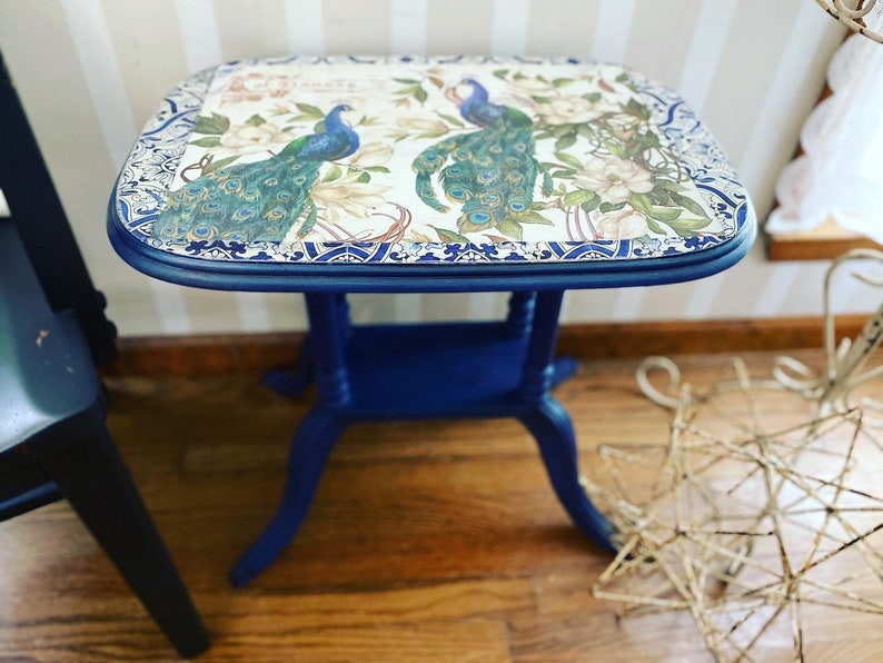 Pode incluir: Uma mesa auxiliar azul com um tampo branco com um design de pav&atilde;o. A mesa tem uma borda decorativa em torno do topo e tr&ecirc;s pernas.