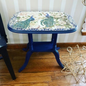 Pode incluir: Uma mesa auxiliar azul com um tampo decorativo com um design de pav&atilde;o. A mesa tem uma borda festonada e um acabamento desgastado.