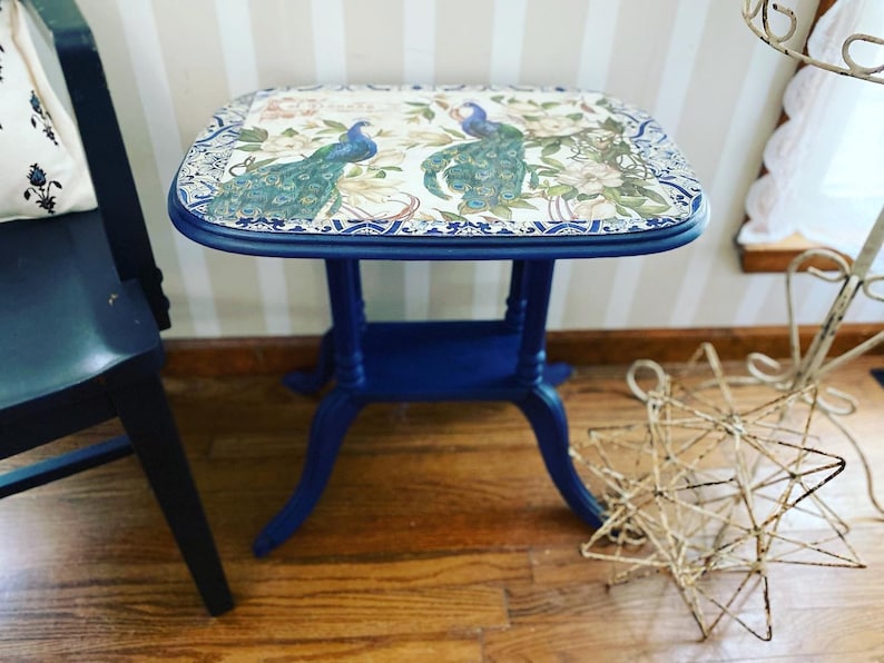 Pode incluir: Uma mesa auxiliar azul com um tampo decorativo com um design de pav&atilde;o. A mesa tem uma borda festonada e um acabamento desgastado.