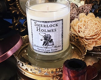 May include: A clear glass candle with a cream-colored wax and a label that reads "Sherlock Holmes." The label features an illustration of Sherlock Holmes smoking a pipe. The candle sits on a gold-colored decorative tray, with a wooden pipe nearby.