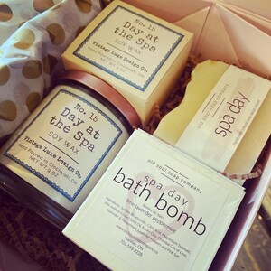 May include: A gift box filled with spa products. The box contains a soy wax candle, a bath bomb, and a bar of soap. The candle and soap are labeled "Day at the Spa" and the bath bomb is labeled "Spa Day" with the scent "Lime Lavender Peppermint".