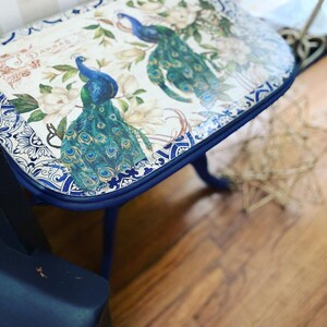 Pode incluir: Uma mesa auxiliar pintada de azul com um tampo estampado em branco e azul com pav&otilde;es. A mesa tem um tampo arredondado e tr&ecirc;s pernas.