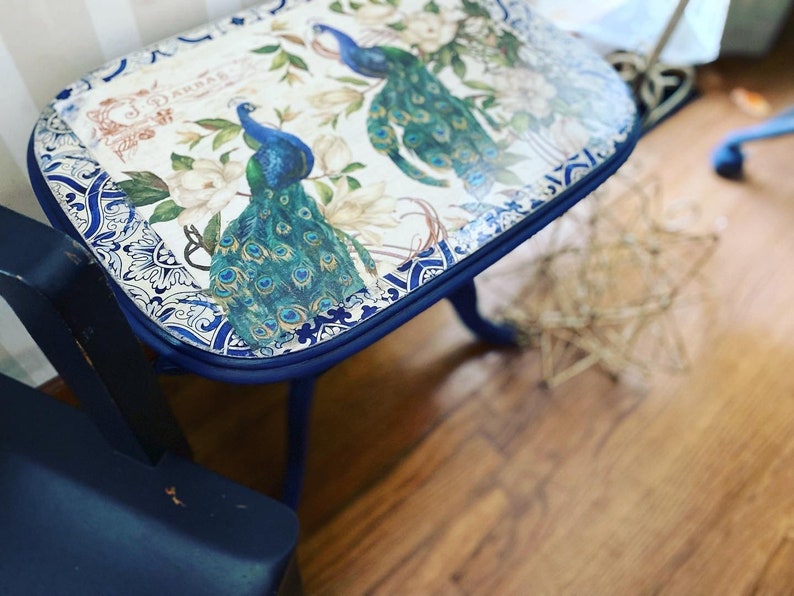 Pode incluir: Uma mesa auxiliar pintada de azul com um tampo estampado em branco e azul com pav&otilde;es. A mesa tem um tampo arredondado e tr&ecirc;s pernas.