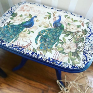 Pode incluir: Uma mesa auxiliar azul com tampo branco com um design de pav&atilde;o e detalhes florais. A mesa tem uma borda decorativa azul e branca.
