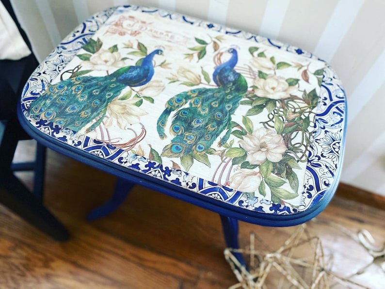 Pode incluir: Uma mesa auxiliar azul com tampo branco com um design de pav&atilde;o e detalhes florais. A mesa tem uma borda decorativa azul e branca.