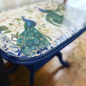 Pode incluir: Uma mesa auxiliar pintada de azul com um tampo branco com um design de pav&atilde;o e um padr&atilde;o floral azul e branco. A mesa tem uma borda arredondada e uma moldura decorativa.