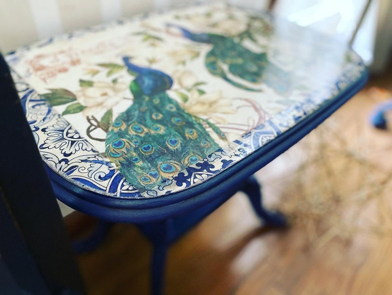 Pode incluir: Uma mesa auxiliar pintada de azul com um tampo branco com um design de pav&atilde;o e um padr&atilde;o floral azul e branco. A mesa tem uma borda arredondada e uma moldura decorativa.