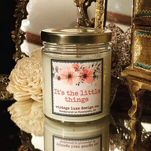 May include: A glass candle jar with a gold lid. The candle is white and has a floral design label with the text "It's the little things vintage luxe design co. Handpoured in Cincinnati, OH."