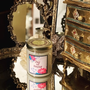 May include: A glass jar candle with a gold lid and a white label that says "Jar of Happy". The label has a floral design with pink, blue, and green colors. The candle is sitting on a gold tray with a mirror surface.