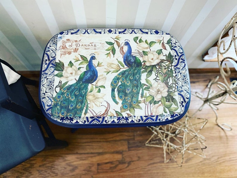 Pode incluir: Uma mesa de madeira com tampo azul e branco com um design decorativo com pav&otilde;es e flores. O design inclui o texto "J. Darbas".