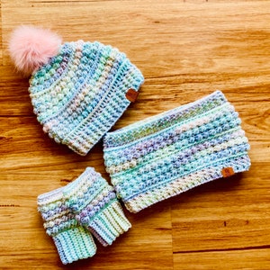 May include: A set of crocheted winter accessories in a pastel colour scheme. The set includes a hat with a fluffy pom-pom, a neck warmer, and fingerless gloves.