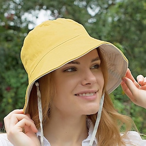 May include: A woman wearing a yellow bucket hat with a white and gray patterned lining. The hat has a chin strap with a black toggle.