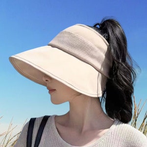 May include: A beige sun visor with a wide brim and a ponytail hole. The visor is worn by a person with long dark hair. The person is standing in front of a beach scene with tall grass.