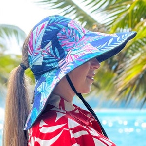 May include: A wide-brimmed sun hat with a tropical leaf print in shades of blue, pink, and green. The hat has a chin strap and a ponytail hole.