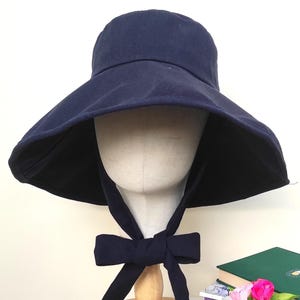 May include: A navy blue sun hat with a wide brim and a matching bow. The hat is displayed on a mannequin head. The hat is on a white table with books and a small vase of flowers.