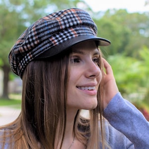 May include: A woman with long brown hair is wearing a blue cardigan and a black and white plaid newsboy cap with a black leather brim. She is smiling and looking to the right.