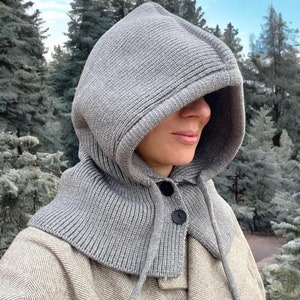 May include: A gray knitted hooded scarf with a button closure. The scarf is worn by a person in a forest setting.