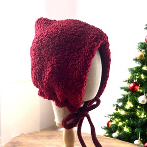 May include: A burgundy knitted baby bonnet with a bow tie. The bonnet is displayed on a wooden stand against a backdrop of a Christmas tree.
