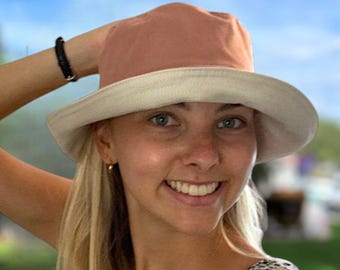 Puede incluir: Una mujer lleva un sombrero de cubo rosa y blanco con un borde blanco. Está sonriendo y mirando a la cámara. El sombrero está hecho de una tela suave y cómoda.