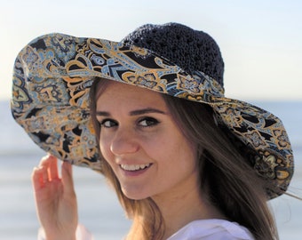 May include: A wide-brimmed sun hat with a dark blue crocheted crown and a patterned brim featuring blue, yellow, and black paisley designs. The hat is worn by a person wearing a white shirt, with a beach and ocean in the background.