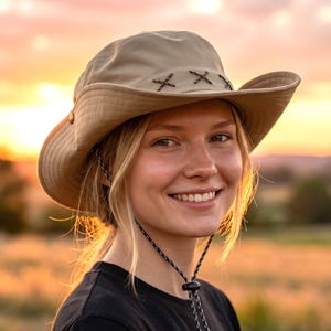 May include: A beige wide-brimmed sun hat featuring a decorative band with three dark brown stitched X-shaped accents. The hat includes a black and white speckled adjustable chin cord with a plastic toggle fastener to secure the fit around the head.