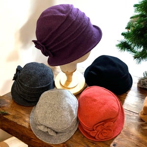 May include: Five felt hats in different colors, including purple, gray, black, and orange. The hats have a wide brim and a flower detail on the side.