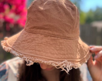 May include: A solid brown bucket hat featuring a textured fabric construction and a wide, downward-sloping brim. The edge of the brim is finished with a frayed, distressed fringe detail that adds a raw, unfinished aesthetic to the design.