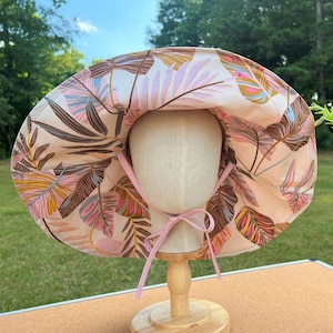 May include: A pink and brown floral sun hat with a wide brim and a pink ribbon tied at the back. The hat is displayed on a wooden stand.