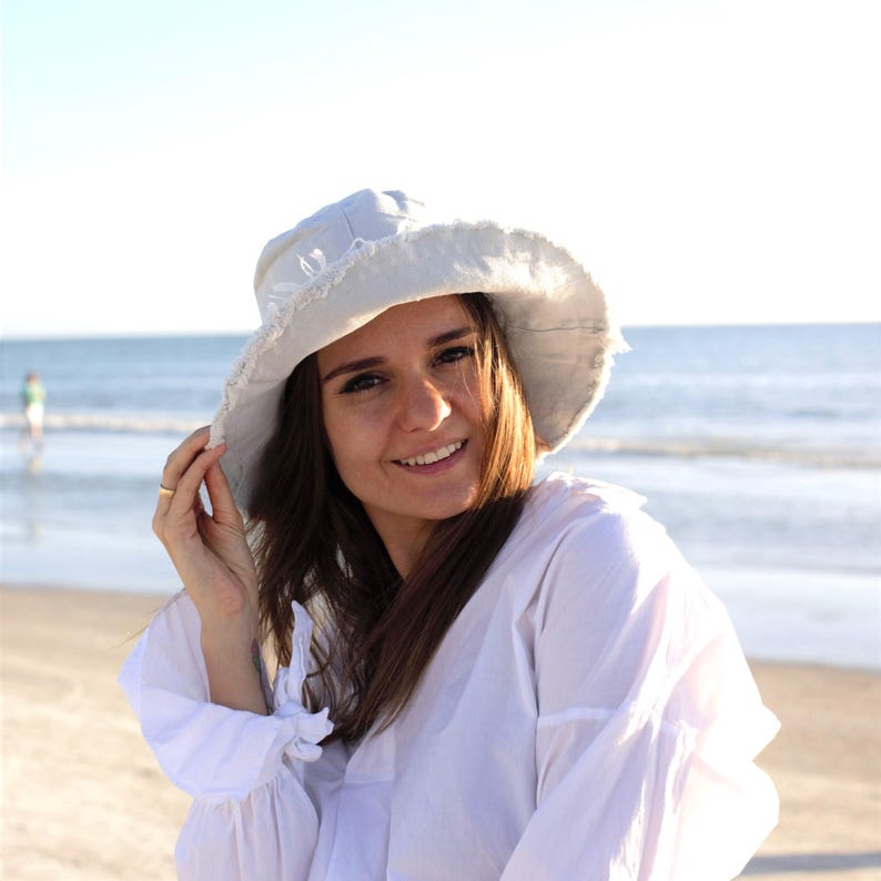 May include: A woman wearing a white shirt and a light gray bucket hat with a frayed edge. She is standing on a beach with the ocean in the background.